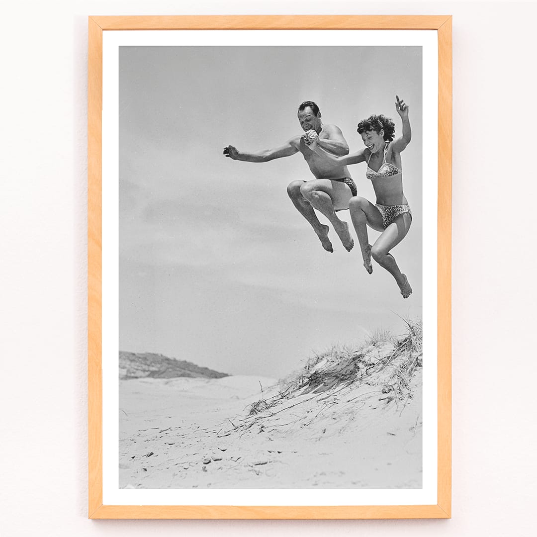 Two Tivoli Stars performing an acrobatic jump on a sandy beach in swimsuits Acrobatics at the Beach poster, framed and displayed in modern living room decor