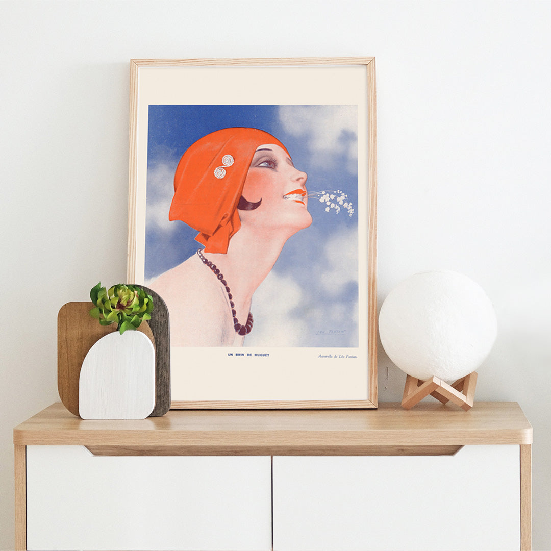 Poster of Un Brin de Muguet by Leo Fontan showing a smiling woman in a red headscarf holding lily of the valley in her mouth, framed and displayed in home decor